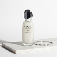 Vixen shimmer oil bottle with geometric cap on a white surface, next to a bracelet.